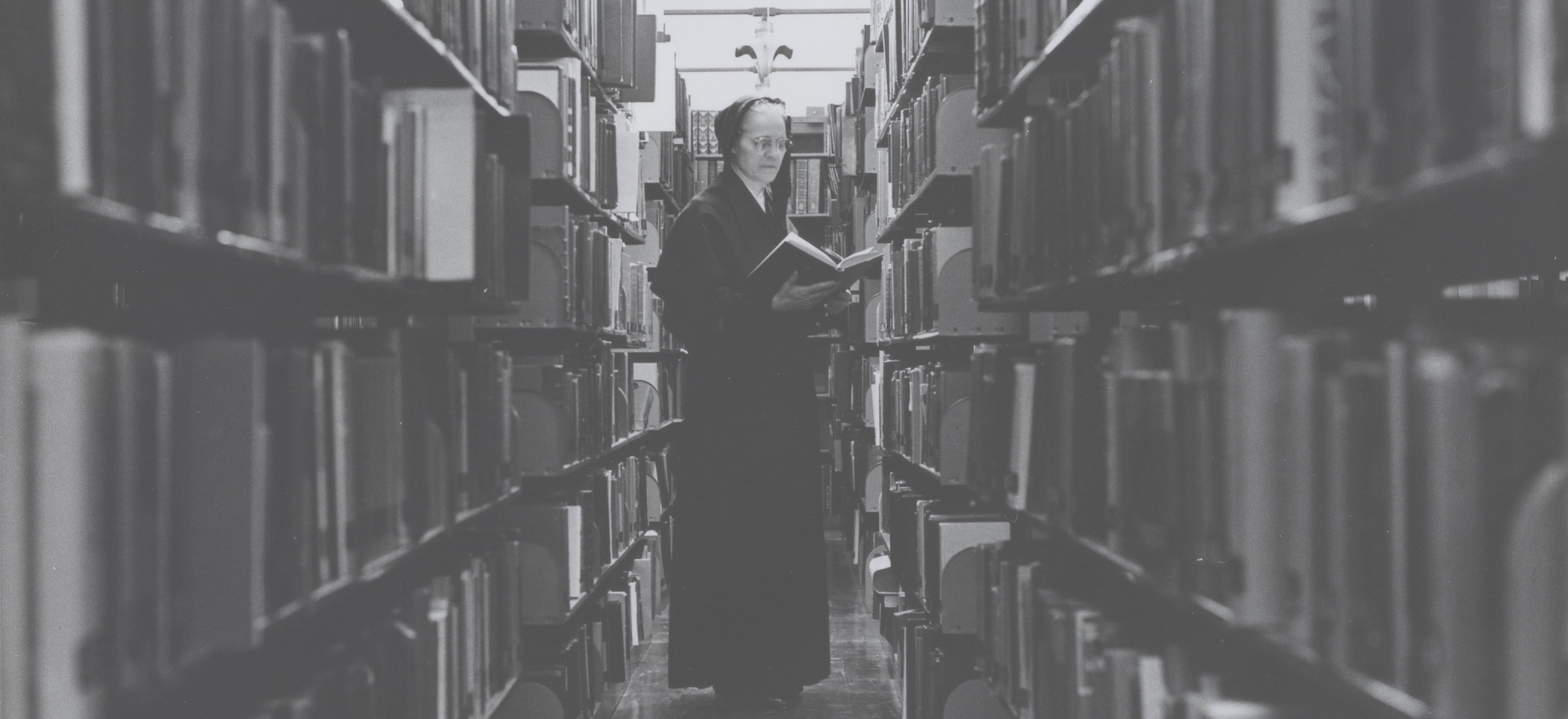A sister wearing a habit is reading a book while standing in an aisle between bookshelves.