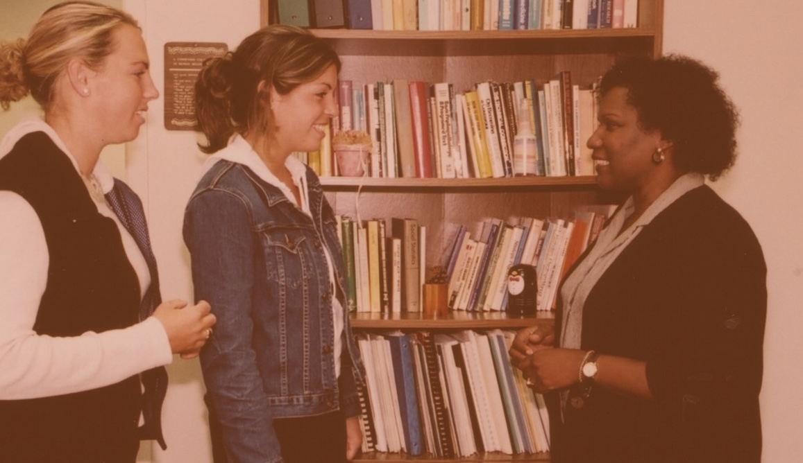 Two students talking to a professor in front of a bookshelf.