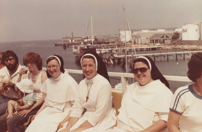Three sisters sitting on a boat in a body of water. There are piers and buildings in the background. People are sitting on either side of the sisters.