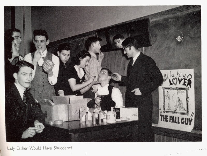 A classroom scene where several people are gathered around a seated individual having makeup applied, with a cluttered table and a poster in the background reading "Will he be a LOVER or THE FALL GUY."