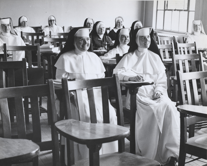 Several sisters in habits are sitting in classroom desks looking at the front of the room.