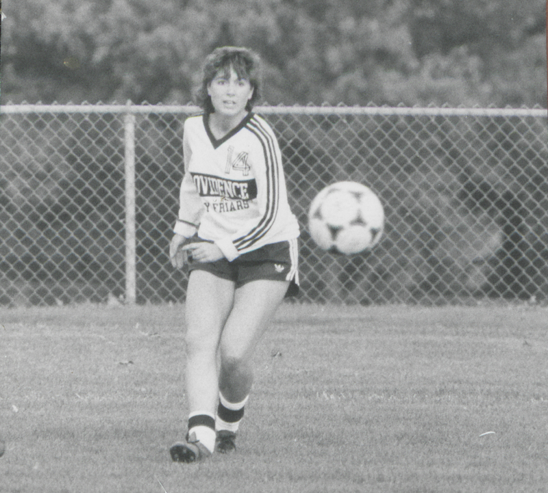 A soccer player in a Providence College uniform runs on a field with a soccer ball in the air near them.