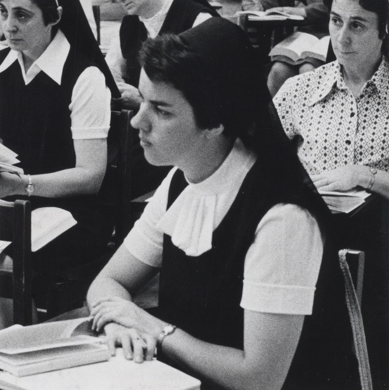 A nun sits at a desk in a classroom with a book in front of her. There are two other nuns sitting behind her.