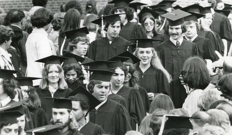 Several students wearing graduation regalia walk down an aisle outdoors as audience members stand on either side of the aisle.
