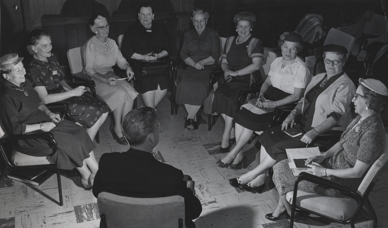 Several women sit in chairs organized into a semi circle in front of a man wearing a clerical suit.
