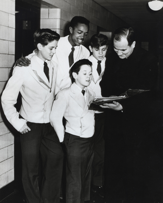 A group of four young boys in school uniforms standing with a reverend. The boys are wearing light-colored blazers, dark ties, and dark pants. The adult man is dressed in a clerical suit and is holding a book or document that the group appears to be looking at together.