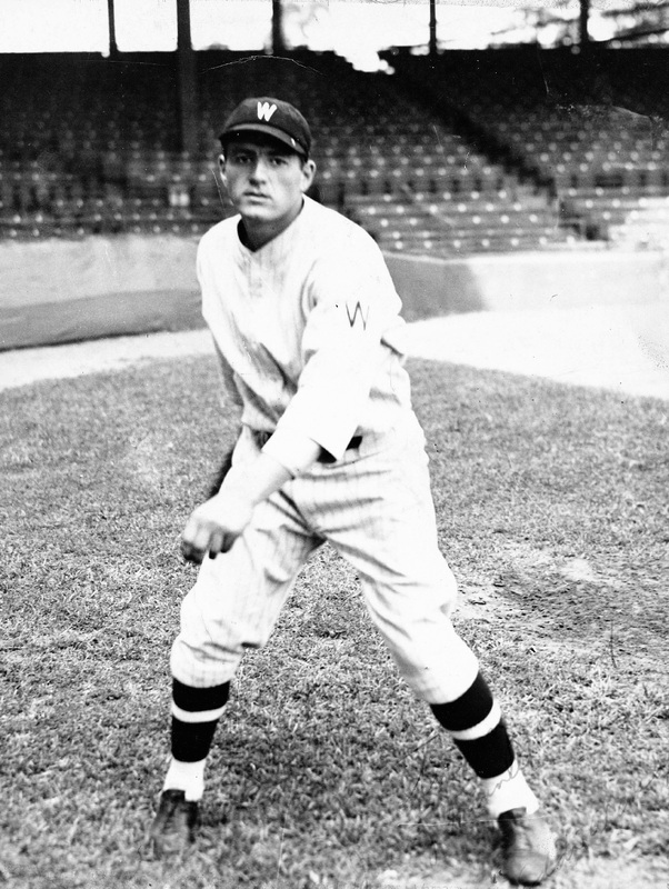 Eddie Wineapple in a pitching stance on a field, wearing a baseball uniform with striped socks and a cap marked with 'W,' with empty stadium seats in the background.