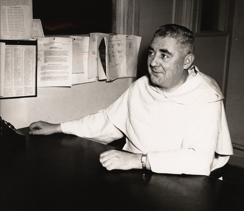 Rev. Vincent Dore in a light-colored robe seated at a desk, with documents pinned to the wall behind them in an office setting.