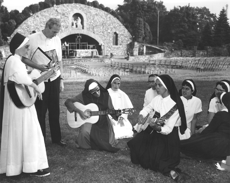 A group of nuns and a man gathered outdoors, playing guitars. In the background, there is an open-air stone structure with an archway and seating area. The nuns are dressed in traditional habits, and some are seated on the grass while others stand.