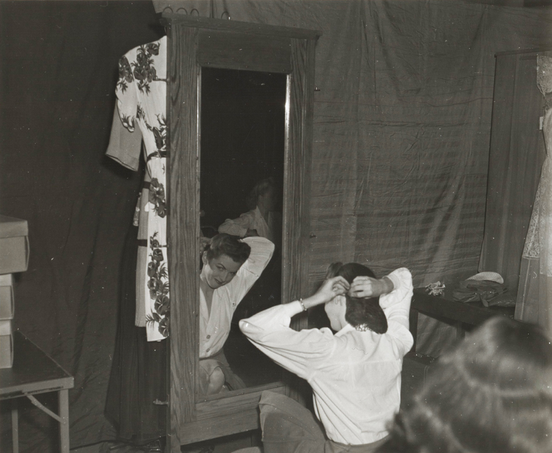 A woman sits on the floor of a dressing room in front of a mirror pinning her hair back.
