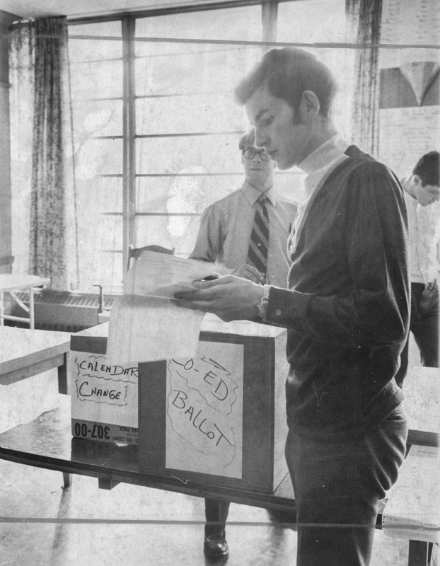 A student standing in front of a ballot box holding a piece of paper. The box reads 'Coed ballot' and 'Calendar change.' There are two other students in the background.