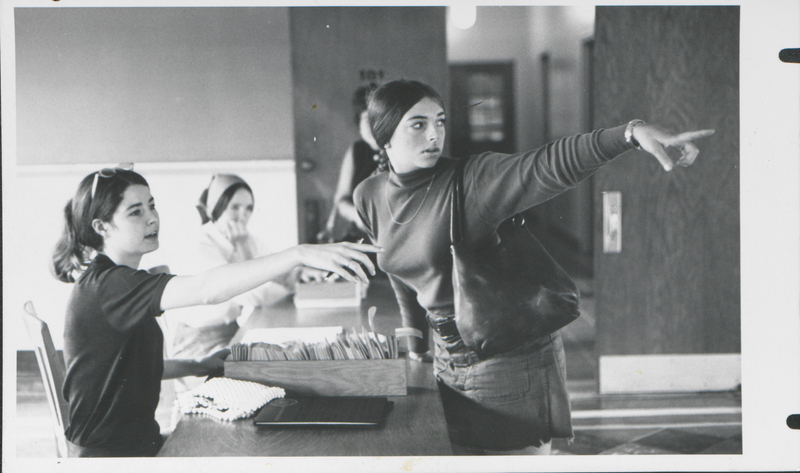 Black and white photograph showing three people in a room. One person is seated at a table with a box of files, reaching out towards another person who is standing and pointing to the right. The third person is partially visible in the background.