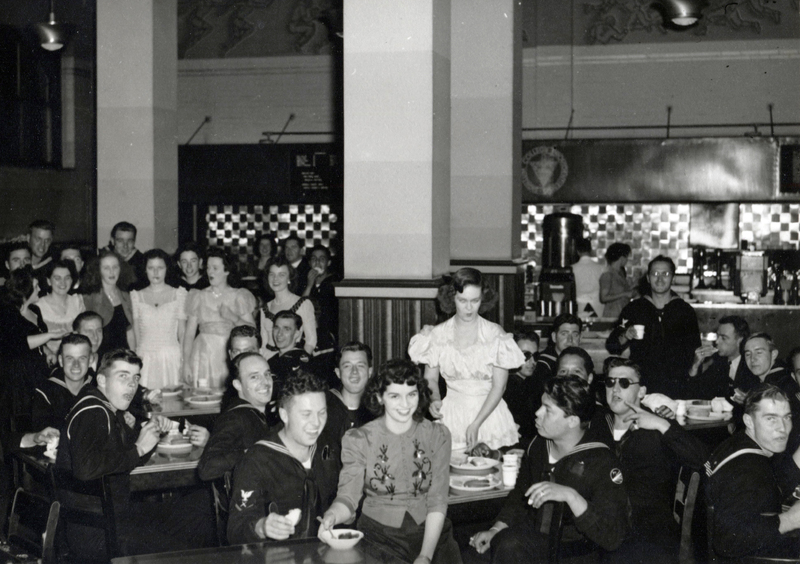 A group of people in a dining area. Some people are seated at tables eating, while others are standing. The setting appears to be a cafeteria or mess hall with several tables and chairs. In the background, there is a counter with food service equipment and tiled walls.
