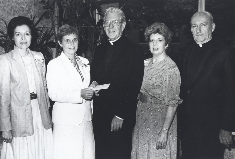 Three women and two men pose for a photograph. One woman is handing one of the men a check. The men are wearing clerical suits.