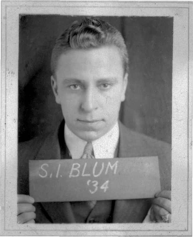 Photo of Samuel Blum in a suit and tie holding a sign that reads "S.I. BLUM '34," looking directly at the camera.
