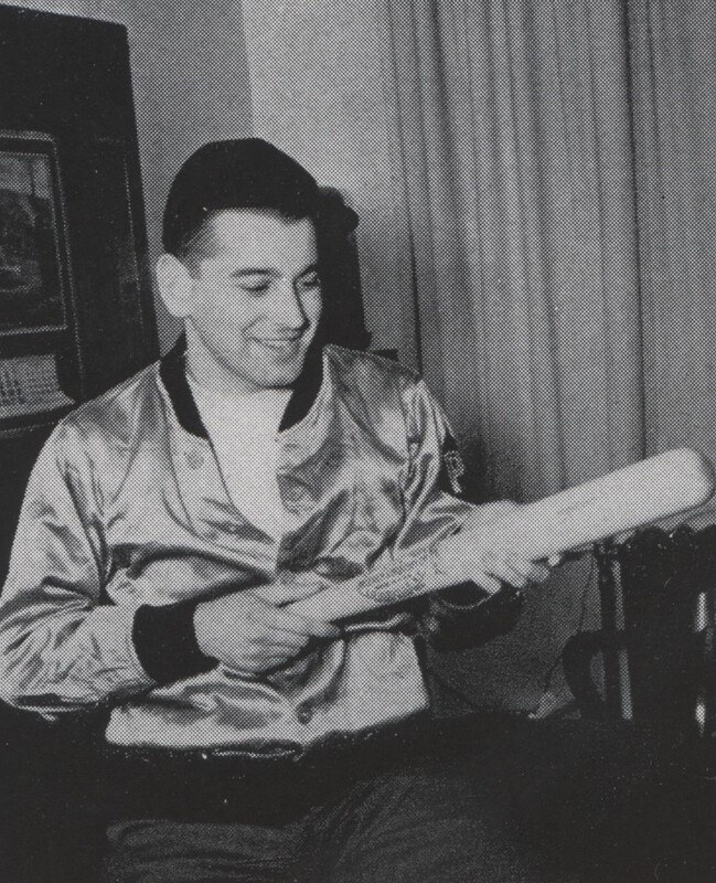 Aaron Slom seated indoors wearing a shiny jacket, holding a baseball bat and looking at it, with a framed picture and curtains visible in the background.