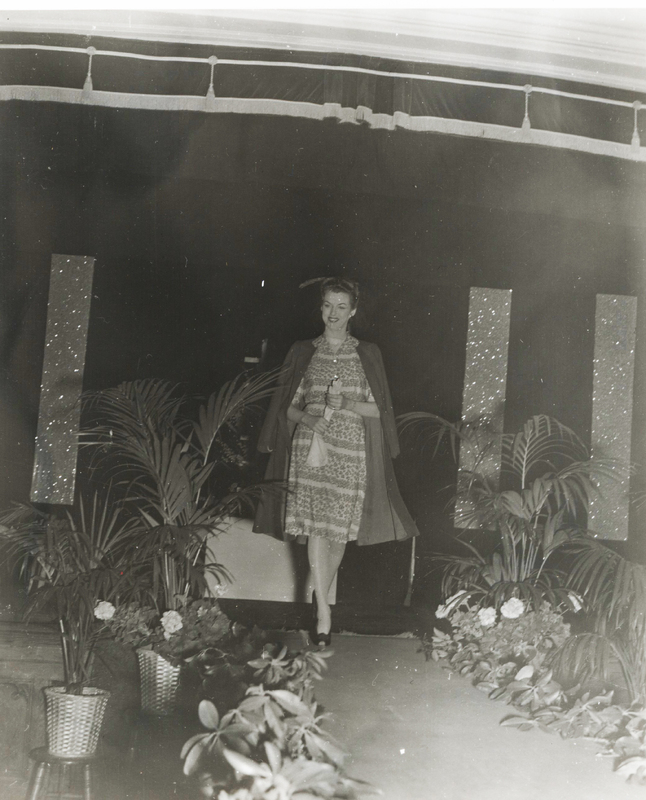 A woman wearing a dress and coat walks down a run way lined with plants.