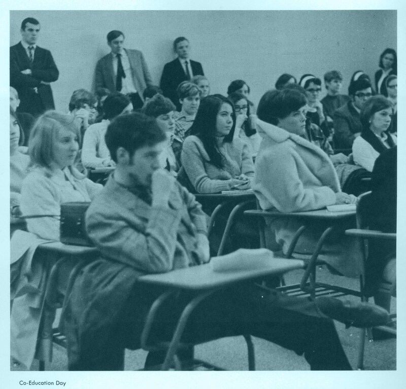 A classroom filled with students most of whom are seated at desks, some taking notes while others listen attentively. The image is labeled 'Co-Education Day' at the bottom left corner.