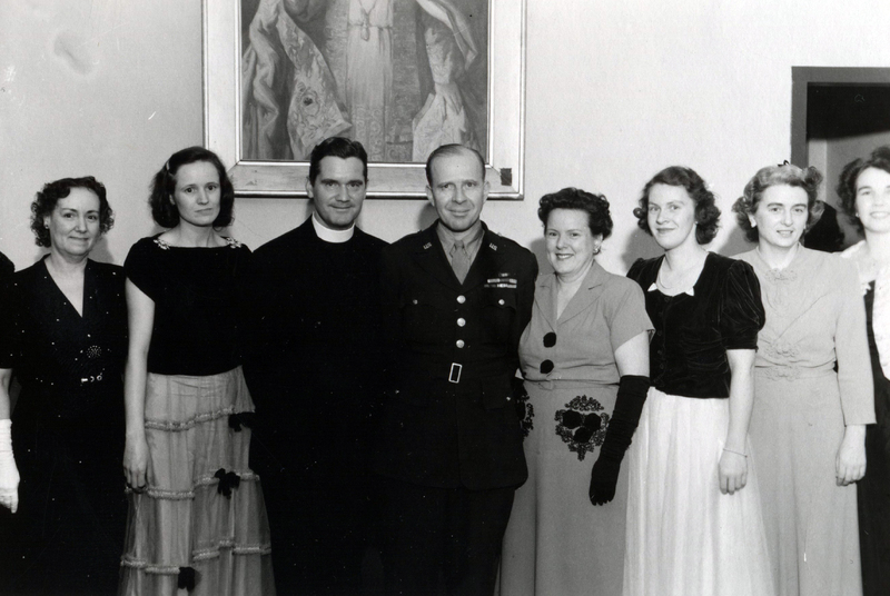 Six women and two men pose for a photo. The women are dressed in gowns and one of the men is in a clerical suit while the other is in a military uniform.