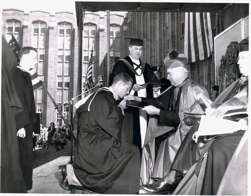 An outdoor formal ceremony with a person in academic regalia kneeling before a seated figure in ceremonial robes, receiving a document, with others in academic attire and American flags in the background.