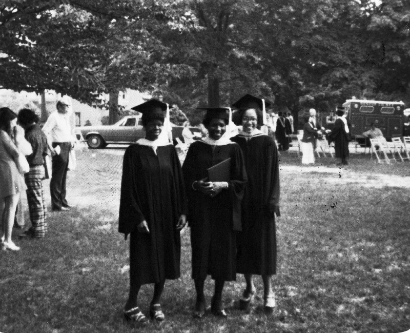 Three women standing on grass outside wearing graduation regalia. One graduate is holding a degree. There are several people in the background and a parked car.