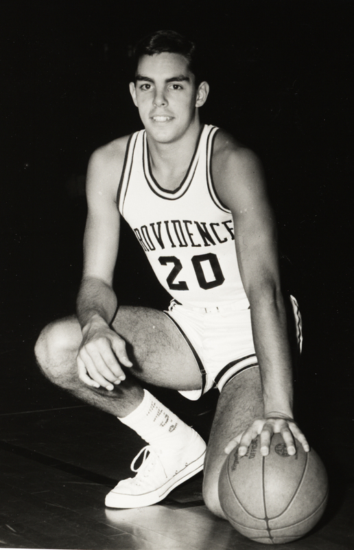 Stuart Kerzner in a PC jersey with the number 20 kneels on one knee, holding a basketball in one hand against a dark background.