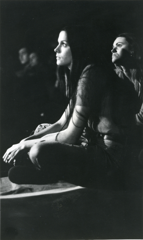 Two women sit on the floor of a dark room looking in one direction. There are some people also sitting on the floor in the background.