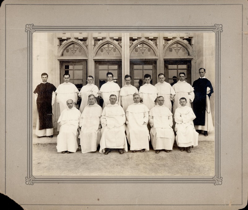 Fourteen clergy members posed in front of a building with Gothic arched windows, with seven seated in white robes and seven standing behind.