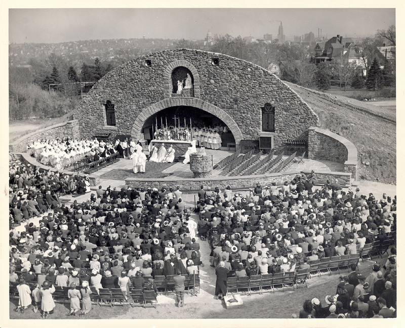 A large outdoor religious ceremony at a stone grotto, with clergy gathered around an altar and a seated crowd in attendance, set against the Providence cityscape.