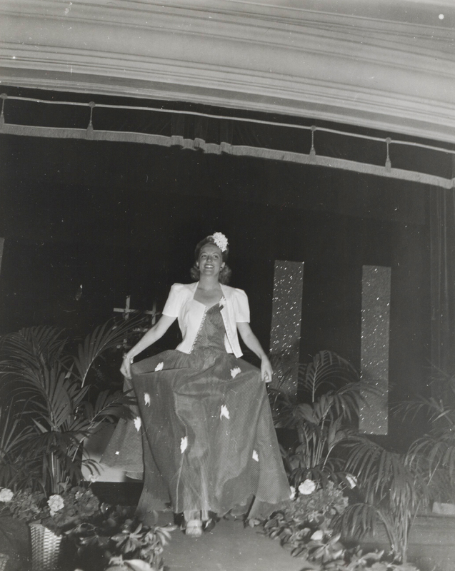 A woman wearing a long dress with a white jacket walks down the runway with plants along the side.