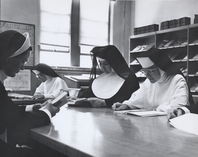 Four nuns are sitting at a table in a library reading from books.
