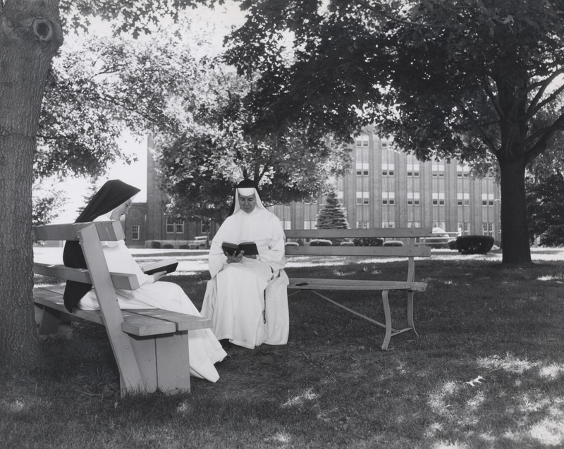 Two nuns are sitting outside on benches below some trees reading from books. There are buildings in the background.