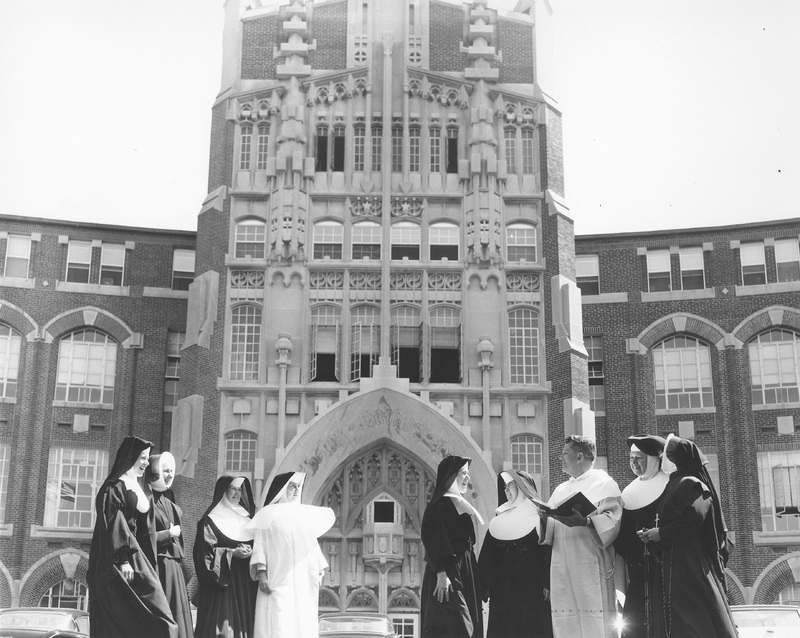 A group of nuns and a priest stand in front of a large, ornate building with Gothic architectural elements. The nuns are dressed in traditional habits, and the priest is wearing clerical robes.