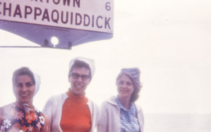 Three women pose outside for a photo standing beneath a sign that partially reads 'Chappaquiddick.'