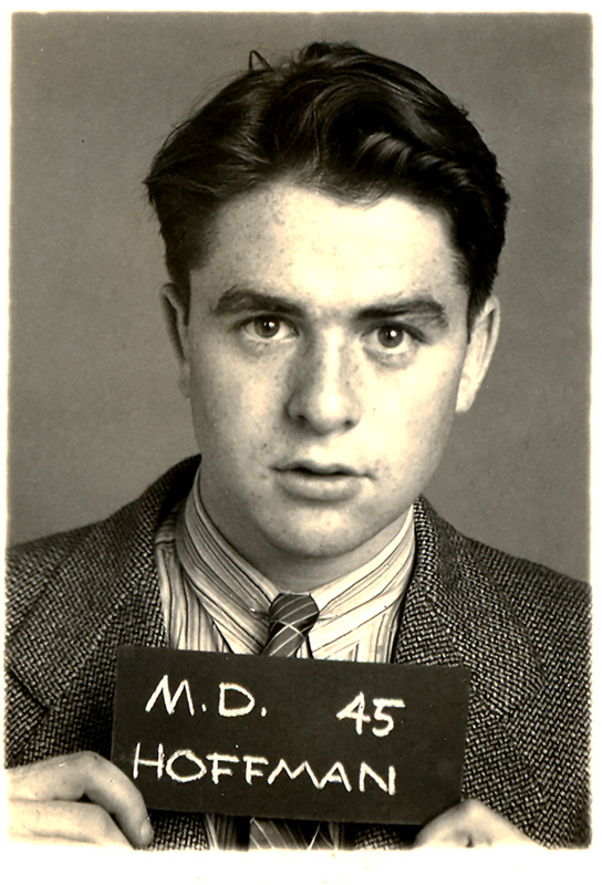 Morton Hoffman in a suit jacket, striped shirt, and tie holding a sign that reads "M.D. 45 HOFFMAN," against a plain background.