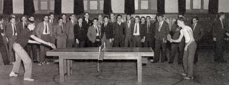 Two people playing table tennis in a large indoor room, with a group of formally dressed spectators watching closely from behind.