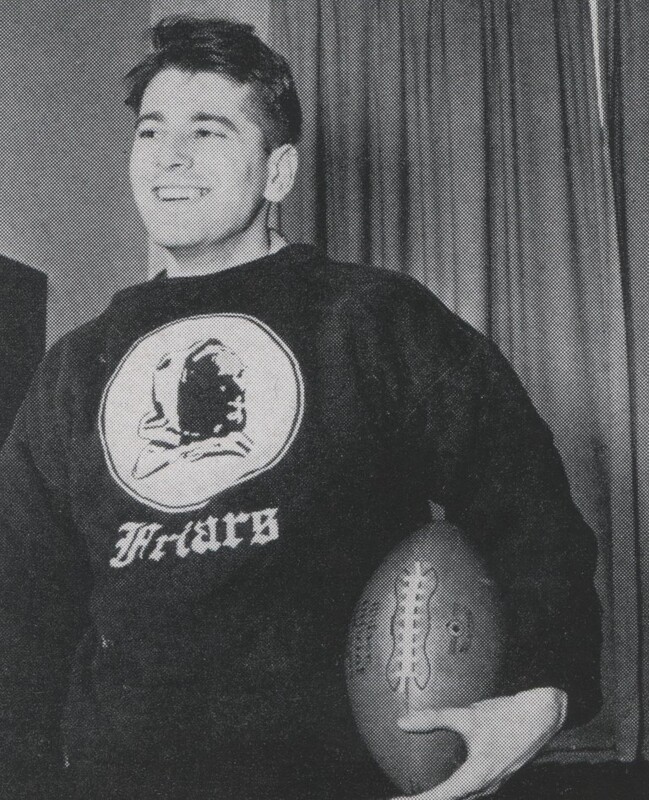 Aaron Slom standing indoors holding a football, wearing a dark sweatshirt with the word "Friars" and an image of a friar on the front, with curtains in the background.