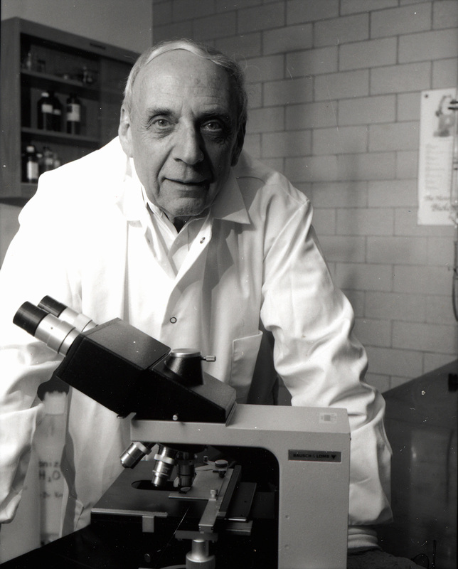 Robert Krasner in a lab coat stands behind a microscope, facing the camera, with a shelf and a brick wall in the background.