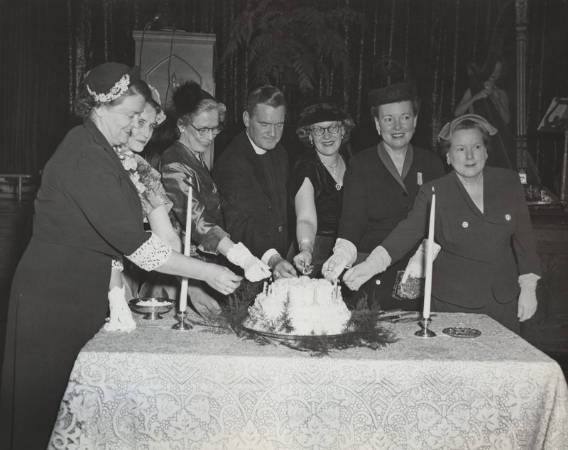 Six women and one man cut into a cake together. The cake sits on a table with a decorative tablecloth and a stage in the background with instruments.