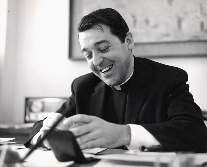 Rev. William Haas in clerical attire seated at a desk, smiling while looking down at an object held in both hands.