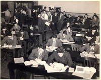 A crowded classroom with students in formal attire seated at desks, reading or writing, suggesting an exam or study session.