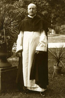 Father James Meagher standing outdoors in a garden, wearing a long white robe with a dark cloak, surrounded by potted plants and trees.