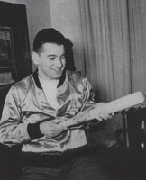 Aaron Slom seated indoors wearing a shiny jacket, holding a baseball bat and looking at it, with a framed picture and curtains visible in the background.