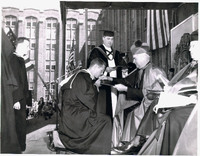 An outdoor formal ceremony with a person in academic regalia kneeling before a seated figure in ceremonial robes, receiving a document, with others in academic attire and American flags in the background.