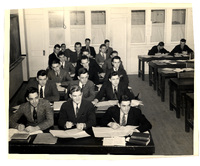 A classroom with students in formal attire seated at desks, reading or writing, with large windows and tables in the background.