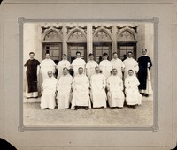 Fourteen clergy members posed in front of a building with Gothic arched windows, with seven seated in white robes and seven standing behind.