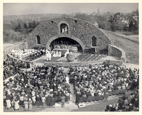 A large outdoor religious ceremony at a stone grotto, with clergy gathered around an altar and a seated crowd in attendance, set against the Providence cityscape.