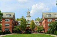 Large, symmetrical brick building with a central archway and clock tower, surrounded by trees and a lawn in the foreground.