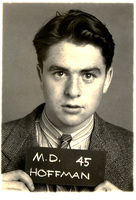 Morton Hoffman in a suit jacket, striped shirt, and tie holding a sign that reads "M.D. 45 HOFFMAN," against a plain background.