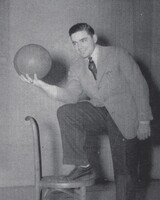 Portrait of Jerome Tesler in a suit posing with one foot on a chair, holding a basketball and smiling at the camera against a plain background.